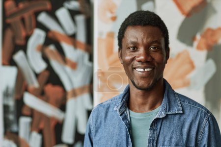 Foto de Joven hombre negro creativo alegre en camisa de mezclilla mirando a la cámara mientras está de pie contra dos pinturas abstractas en el estudio de las artes - Imagen libre de derechos