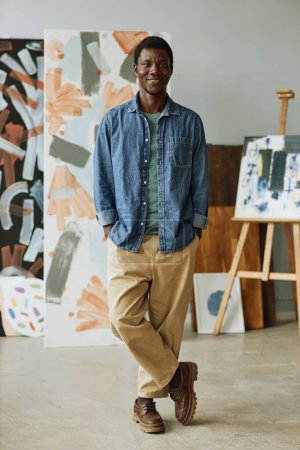 Téléchargez les photos : Joyeux jeune artisan créatif en tenue décontractée debout devant la caméra en studio d'art contre la collecte de ses peintures - en image libre de droit
