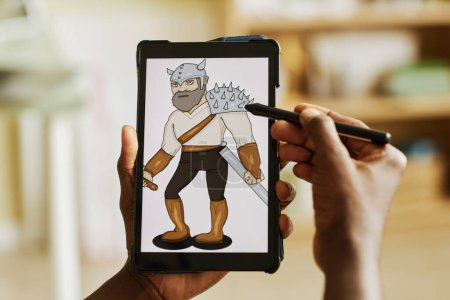 Foto de Tableta con imagen gráfica del guerrero en la pantalla en manos del diseñador afroamericano usando lápiz óptico mientras crea una nueva imagen - Imagen libre de derechos