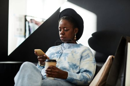 Foto de Retrato gráfico de una elegante mujer negra que usa sombrero mientras se relaja en el salón del centro comercial y usa un teléfono inteligente, espacio para copiar - Imagen libre de derechos