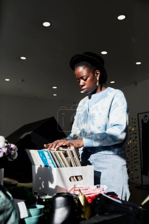 Téléchargez les photos : Portrait vertical d'une jeune femme noire regardant des disques vintage dans un magasin éclairé par la lumière du soleil - en image libre de droit
