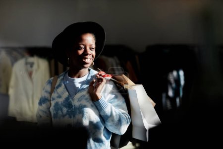 Foto de Retrato de moda de una mujer negra sonriente sosteniendo bolsas de compras en una tienda de segunda mano iluminada por la luz del sol, espacio para copiar - Imagen libre de derechos