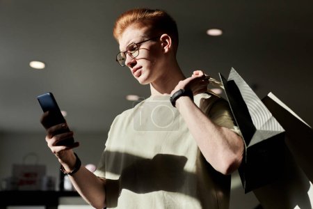Téléchargez les photos : Portrait latéral du jeune homme de la génération Z utilisant un téléphone intelligent tout en appréciant les achats au magasin d'épargne éclairé par la lumière du soleil - en image libre de droit