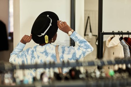 Foto de Elegante mujer negra probándose el sombrero en la tienda de segunda mano y mirando en el espejo, etiqueta de la venta, espacio de copia - Imagen libre de derechos