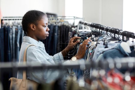 Foto de Retrato de vista lateral de una joven mujer negra mirando la ropa en un estante en una tienda de segunda mano - Imagen libre de derechos