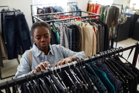 Téléchargez les photos : Portrait grand angle de jeune femme noire regardant des vêtements tout en faisant des emplettes pour des affaires dans le magasin d'épargne - en image libre de droit