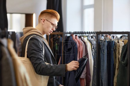 Foto de Retrato de vista lateral de un joven mirando ropa mientras hace compras sostenibles en una tienda de segunda mano - Imagen libre de derechos