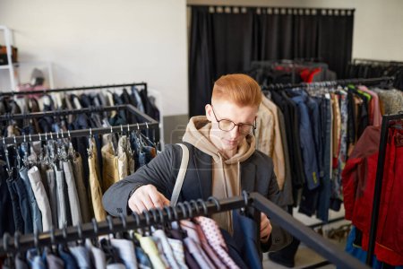 Foto de Retrato de alto ángulo del joven pelirrojo mirando la ropa mientras hace compras sostenibles en una tienda de segunda mano - Imagen libre de derechos
