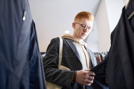 Photo for Low angle portrait of red haired young man browsing clothes on racks while shopping sustainably in thrift store - Royalty Free Image