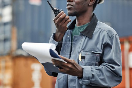 Photo for Cropped shot of young black man using walkie talkie while working at shipping docks with containers - Royalty Free Image
