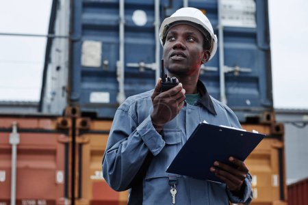 Photo for Waist up portrait of black male worker wearing hardhat and talking to radio in shipping docks with containers, copy space - Royalty Free Image