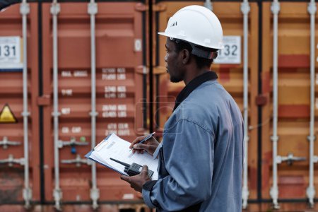 Photo for Side view portrait of young male worker wearing writing on clipboard in shipping docks with containers, copy space - Royalty Free Image