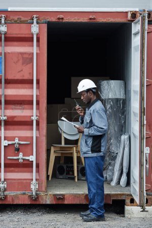 Téléchargez les photos : Portrait vertical latéral du jeune travailleur portant un casque de sécurité lors de la vérification des conteneurs au quai d'expédition - en image libre de droit