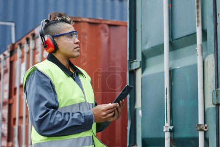 Photo for Side view portrait of female worker wearing ear protection and holding tablet while checking containers at shipping docks, copy space - Royalty Free Image