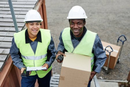 Photo for High angle view at two smiling workers looking at camera outdoors while carrying boxes up stairs at shipping docks - Royalty Free Image