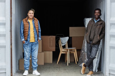 Photo for Full length portrait of two smiling young people standing inside shipping container with boxes in background, copy space - Royalty Free Image