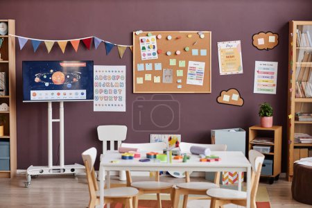 Téléchargez les photos : Intérieur de salle de classe spacieuse pour les enfants d'âge préscolaire avec table entourée de chaises au centre et des conseils avec des notes et des photos - en image libre de droit