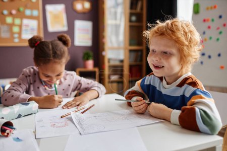 Téléchargez les photos : Joyeux apprenant mignon de la maternelle avec des taches de rousseur et des cheveux roux tenant crayon tout en étant assis à table contre camarade de classe et dessin - en image libre de droit