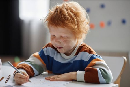 Photo for Cute primary school learner with freckles and ginger hair drawing with pencil while sitting by table in front of camera at lesson - Royalty Free Image