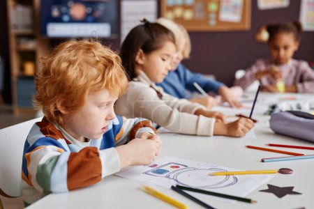 Téléchargez les photos : Rangée d'apprenants interculturels assidus de l'école maternelle assis à table et dessinant avec des crayons avec un mignon garçon aux cheveux roux devant - en image libre de droit