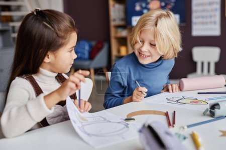 Téléchargez les photos : Petit garçon blond regardant une image dessinée et tenue par son camarade de classe lui montrant à la leçon de dessin à l'école maternelle - en image libre de droit