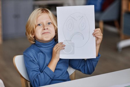 Téléchargez les photos : Mignon blond garçon imitant expression de sombre visage dessiné sur papier qu'il vous montre tout en étant assis à table à la leçon de dessin - en image libre de droit