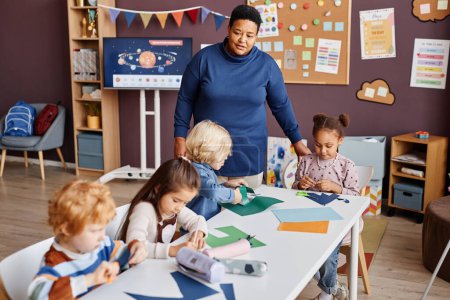 Téléchargez les photos : Professeur confiant debout devant la table où groupe de petits apprenants de l'école primaire créant des jouets en papier ou origami à la leçon - en image libre de droit