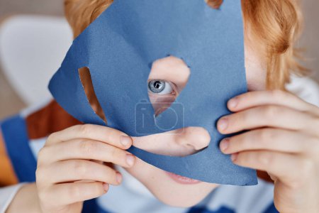 Téléchargez les photos : Gros plan de mignon petit garçon aux yeux bleus couvrant le visage avec un masque en papier bleu fait à la main tout en gardant les yeux et le nez dans les trous coupés - en image libre de droit