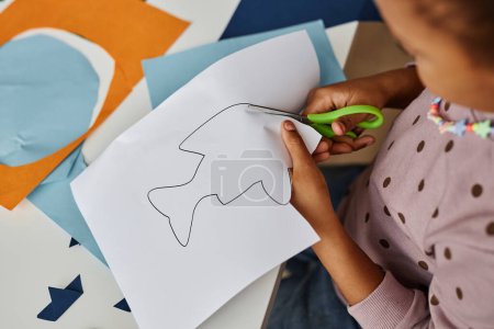 Foto de Por encima de tiro de niña afroamericana cortando el contorno de pescado dibujado en el papel blanco mientras se crean juguetes en la lección en la escuela infantil - Imagen libre de derechos