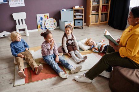 Photo for Group of curious intercultural kids relaxing on carpet in front of teacher reading them fairy tale in the miidle of the day at nursery school - Royalty Free Image