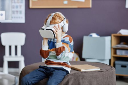 Photo for Gingerhaired little boy in striped pullover and vr headset playing virtual game while sitting on round soft ottoman in nursery school - Royalty Free Image