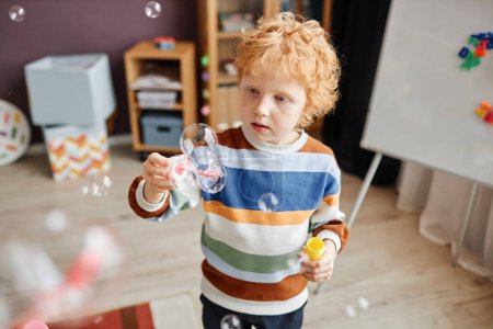 Photo for Little boy with short ginger hair in striped pullover looking at multiple soap bubble while playing in front of camera in kindergarten - Royalty Free Image