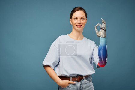 Photo for Cheerful young woman with bionic arm showing ok sign, isolated on greyish blu - Royalty Free Image