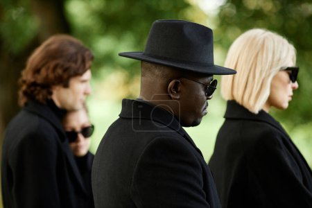 Foto de Vista lateral de personas vestidas de negro con gafas de sol de pie en silencio en la ceremonia funeraria al aire libre - Imagen libre de derechos