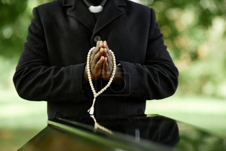 Foto de Primer plano del sacerdote afroamericano vestido de negro en la ceremonia funeraria al aire libre con enfoque en las manos en la oración - Imagen libre de derechos