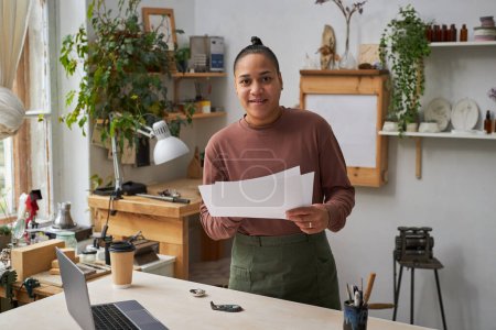 Foto de Retrato de artista femenina multiétnica sonriendo a la cámara mientras trabaja en nuevos diseños en el estudio - Imagen libre de derechos