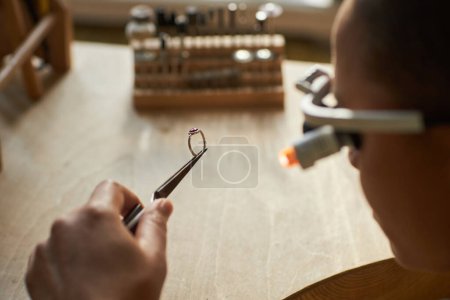Foto de Macro shot de joyero artesanal sosteniendo anillo hecho a mano con piedra rubí mientras se crean piezas de arte sobre mesa de madera, espacio de copia - Imagen libre de derechos