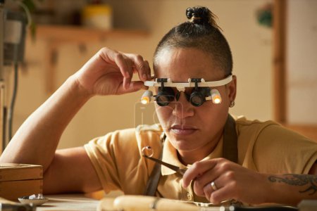 Foto de Joyero femenino sosteniendo anillo hecho a mano y usando gafas de aumento en acogedor estudio - Imagen libre de derechos