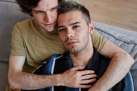 Téléchargez les photos : Portrait à angle élevé de couple gay embrassant à la maison avec un accent sur jeune homme regardant la caméra - en image libre de droit