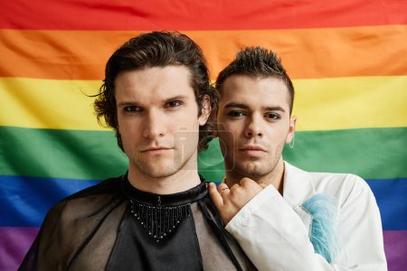 Foto de Retrato de dos jóvenes gay mirando a la cámara con la bandera del orgullo LGBTQ en el fondo - Imagen libre de derechos