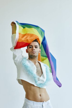 Téléchargez les photos : Portrait vertical d'un jeune homme gay agitant son drapeau de fierté et portant un haut de culture contre le blanc - en image libre de droit