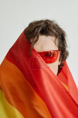 Téléchargez les photos : Concept shot de gay jeune homme enveloppé dans la fierté drapeau, demi-visage, rouge lunettes de soleil - en image libre de droit