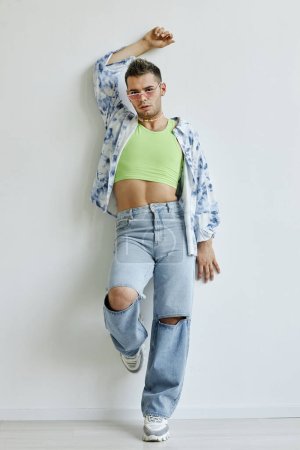 Vertical full length portrait of extravagant young gay man leaning against white wall in fashion studio