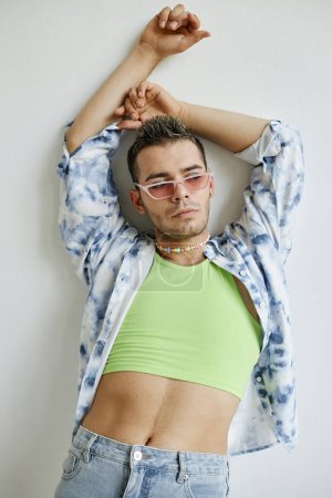 Téléchargez les photos : Portrait vertical de l'homme gay confiant posant sensuellement appuyé contre un mur blanc - en image libre de droit