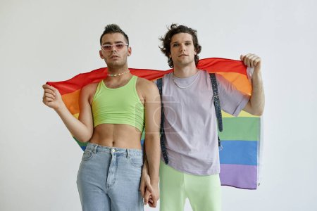 Photo for Portrait of two gay young men holding hands with pride flag against white background - Royalty Free Image
