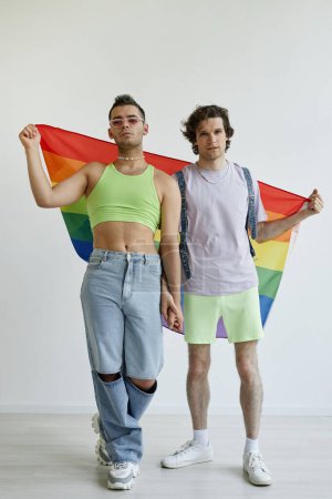 Photo for Full length portrait of two gay men holding hands posing with pride flag against white background - Royalty Free Image