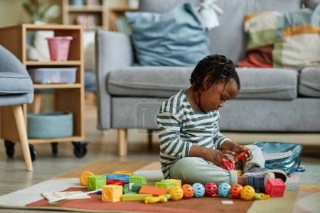 Photo for Side view portrait of cute black baby playing with toys while sitting on floor in cozy home, copy space - Royalty Free Image