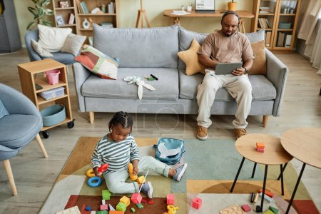 Téléchargez les photos : Vue grand angle au bébé garçon noir jouant avec des jouets sur le sol pendant que le père travaille en arrière-plan, copier l'espace - en image libre de droit