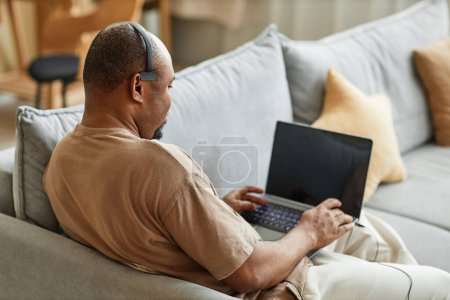 Téléchargez les photos : Vue arrière à l'homme noir adulte en utilisant un ordinateur portable avec casque tout en travaillant dans une réunion en ligne de la maison, espace de copie - en image libre de droit