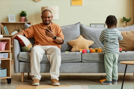 Foto de Retrato completo del padre usando smartphone con hijo jugando en el sofá en casa, espacio para copiar - Imagen libre de derechos